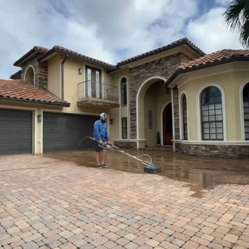 "Professional driveway cleaning in action as a technician uses specialized equipment to thoroughly clean a brick driveway in front of a large, elegant home."